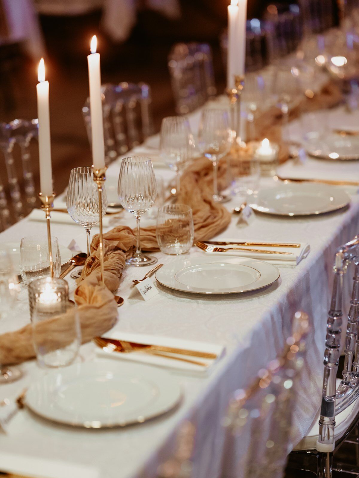A long table set before the guests sit: white linen, gold cutlery, crystal glasses and tall lit candles.
